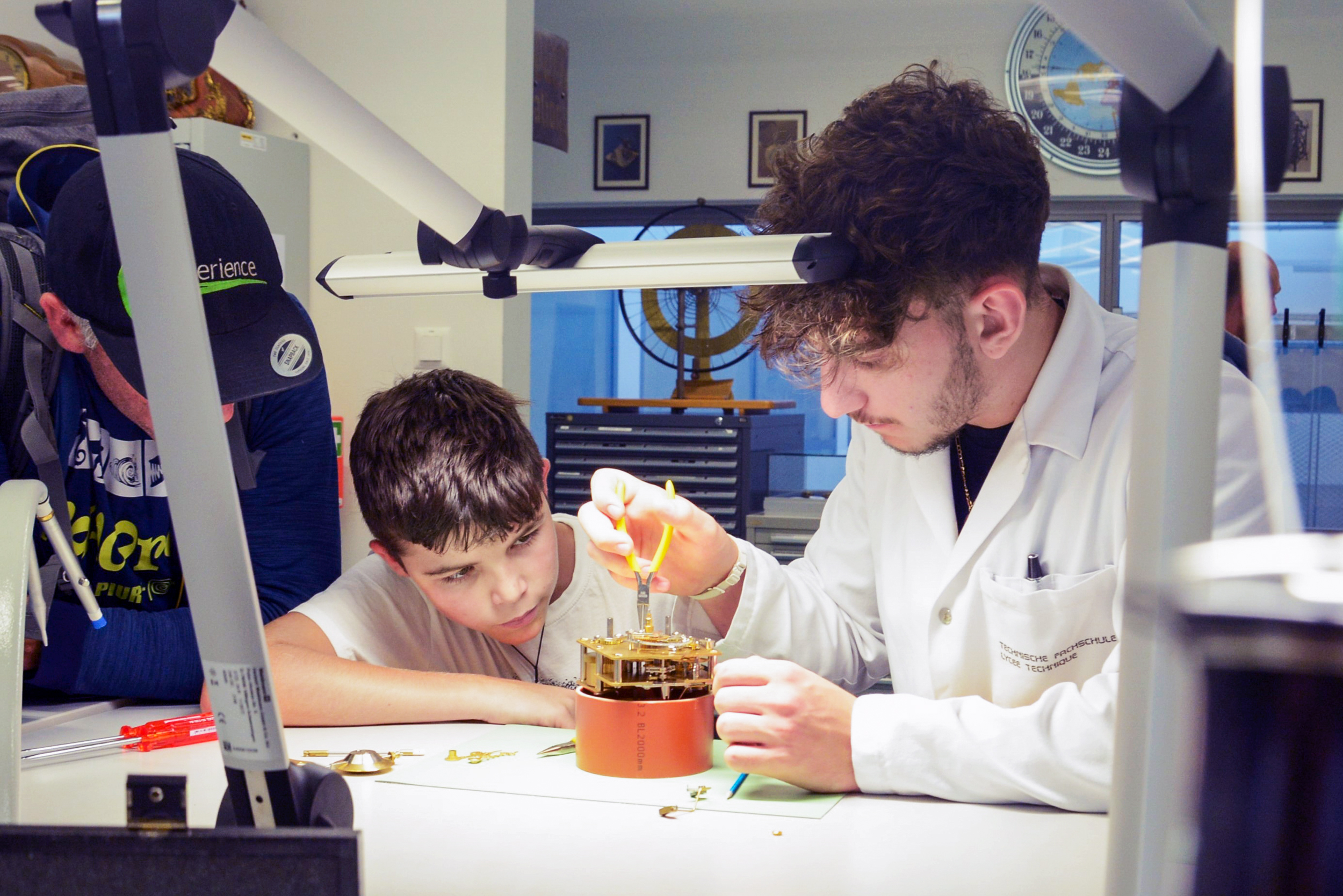 : Lernender zeigt einem Jungen wie er an einem Uhrwerk arbeitet